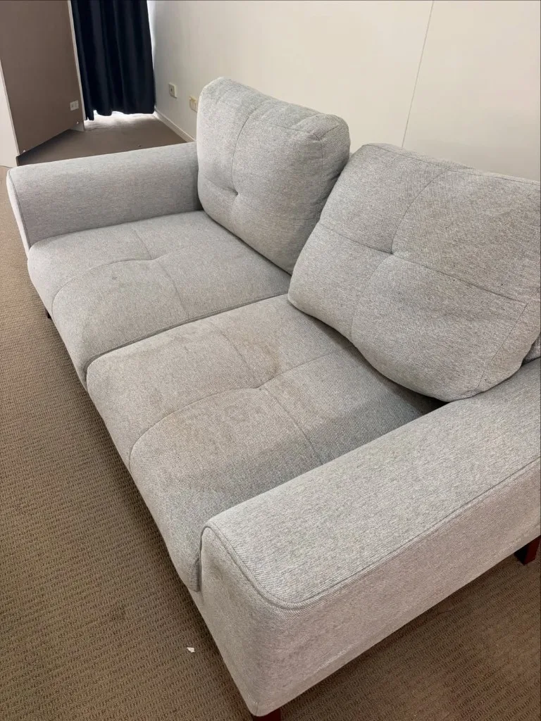 Grey fabric two-seater couch with visible brown stains on the seat cushions before cleaning.
