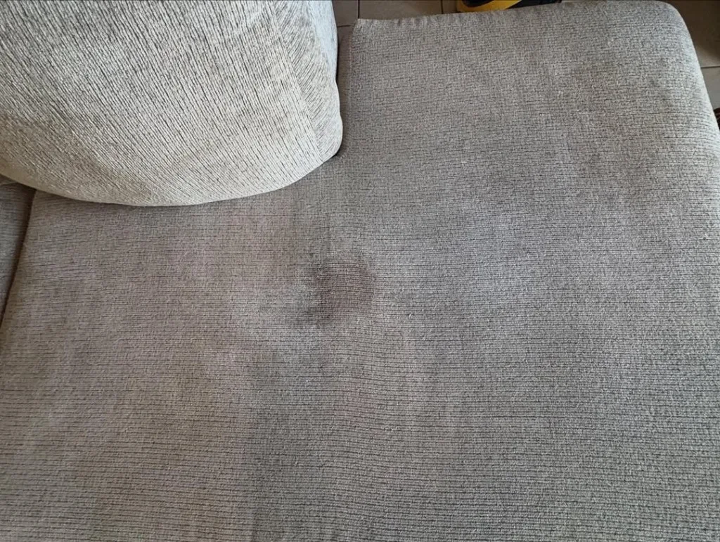 Close-up of a light fabric sofa cushion with a dark stain before cleaning.